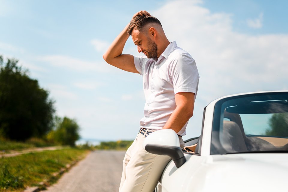 Man standing beside a white car on a quiet road, running his hand through his hair, appearing thoughtful and reflective after a driving experience.