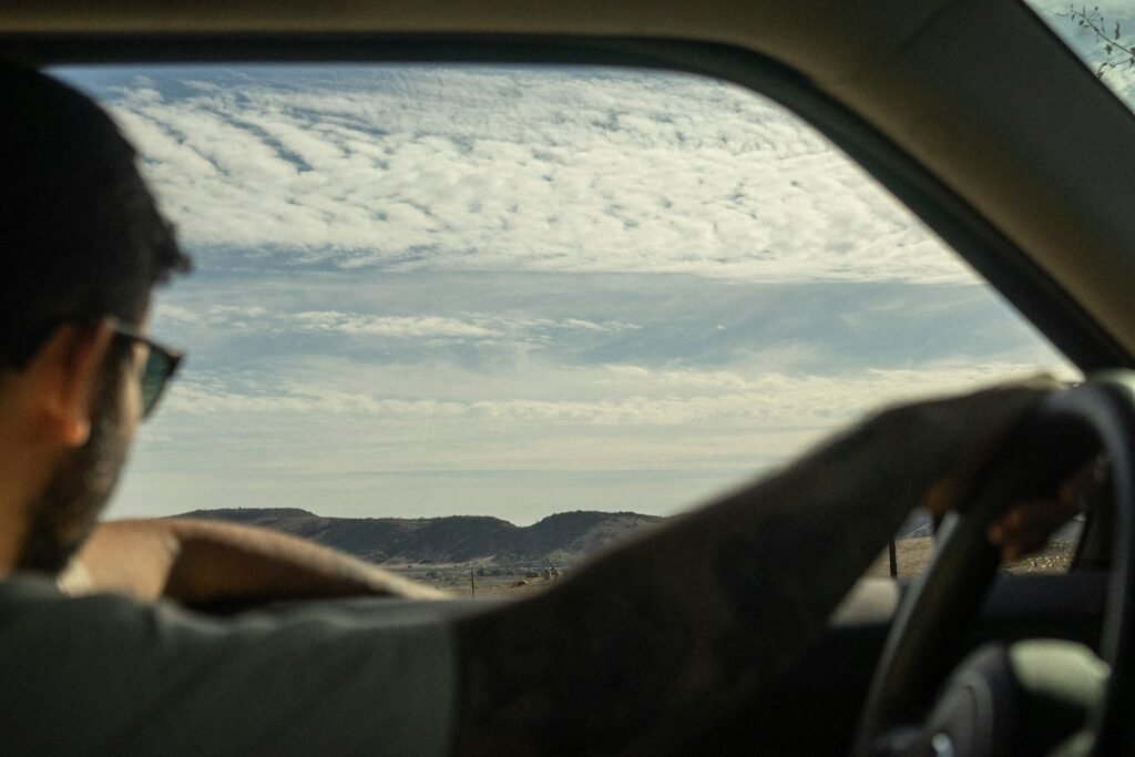 A man wearing sunglasses drives through a scenic desert landscape with hills in the distance, his arm resting casually on the car window as he looks out at the view under a partly cloudy sky.